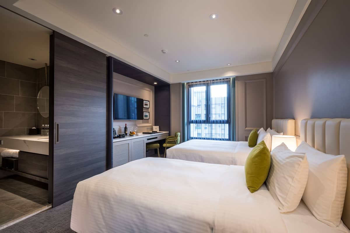 A modern hotel room with two beds, a bathroom, a desk, and a large window.