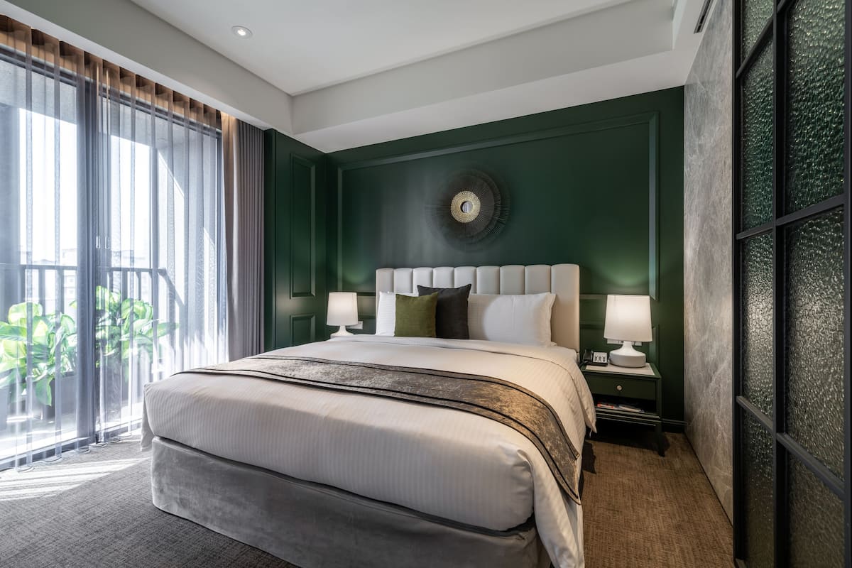 A modern hotel room with a large bed, a nightstand with a lamp, and a view of greenery outside.