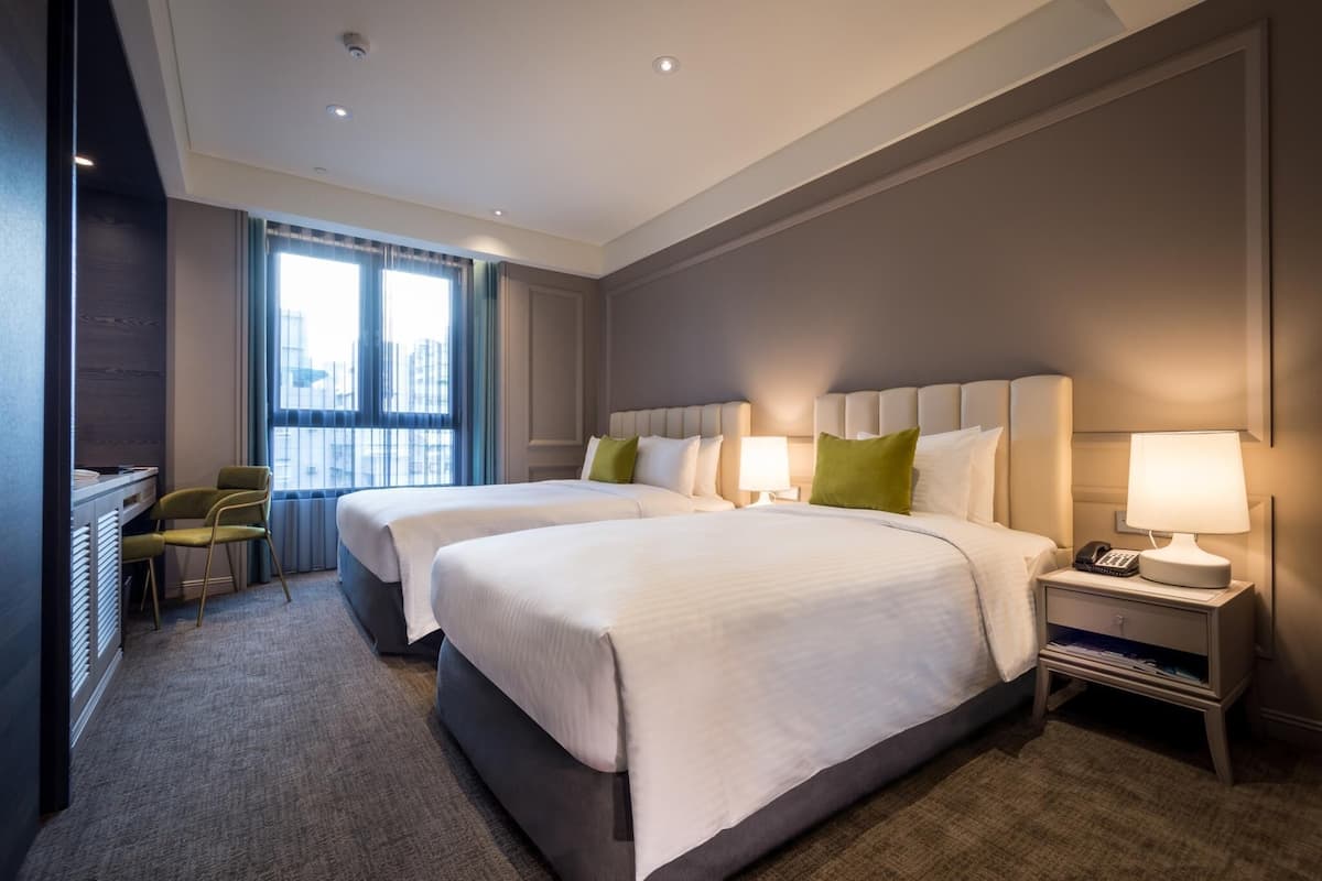 A hotel room with two beds, a desk, a chair, a nightstand with a lamp, and a window with curtains.