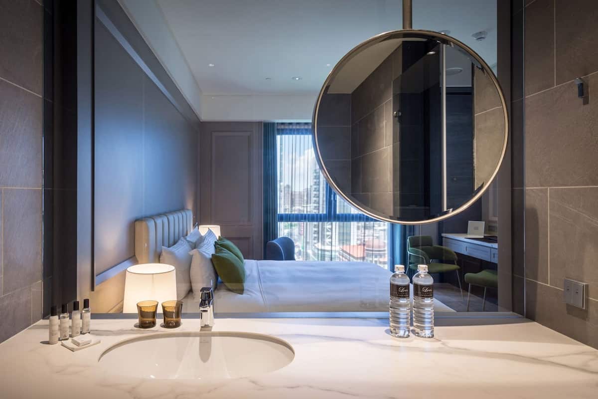 A modern hotel bathroom with a large mirror, a marble sink, toiletries, and a bed with a city view.