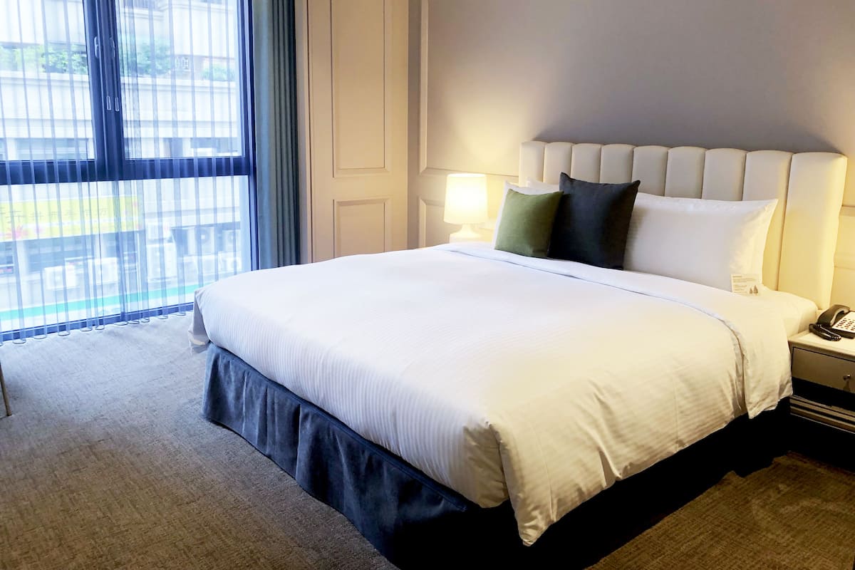 A hotel room with a large bed, a bedside table with a lamp, and a view through a window with curtains.