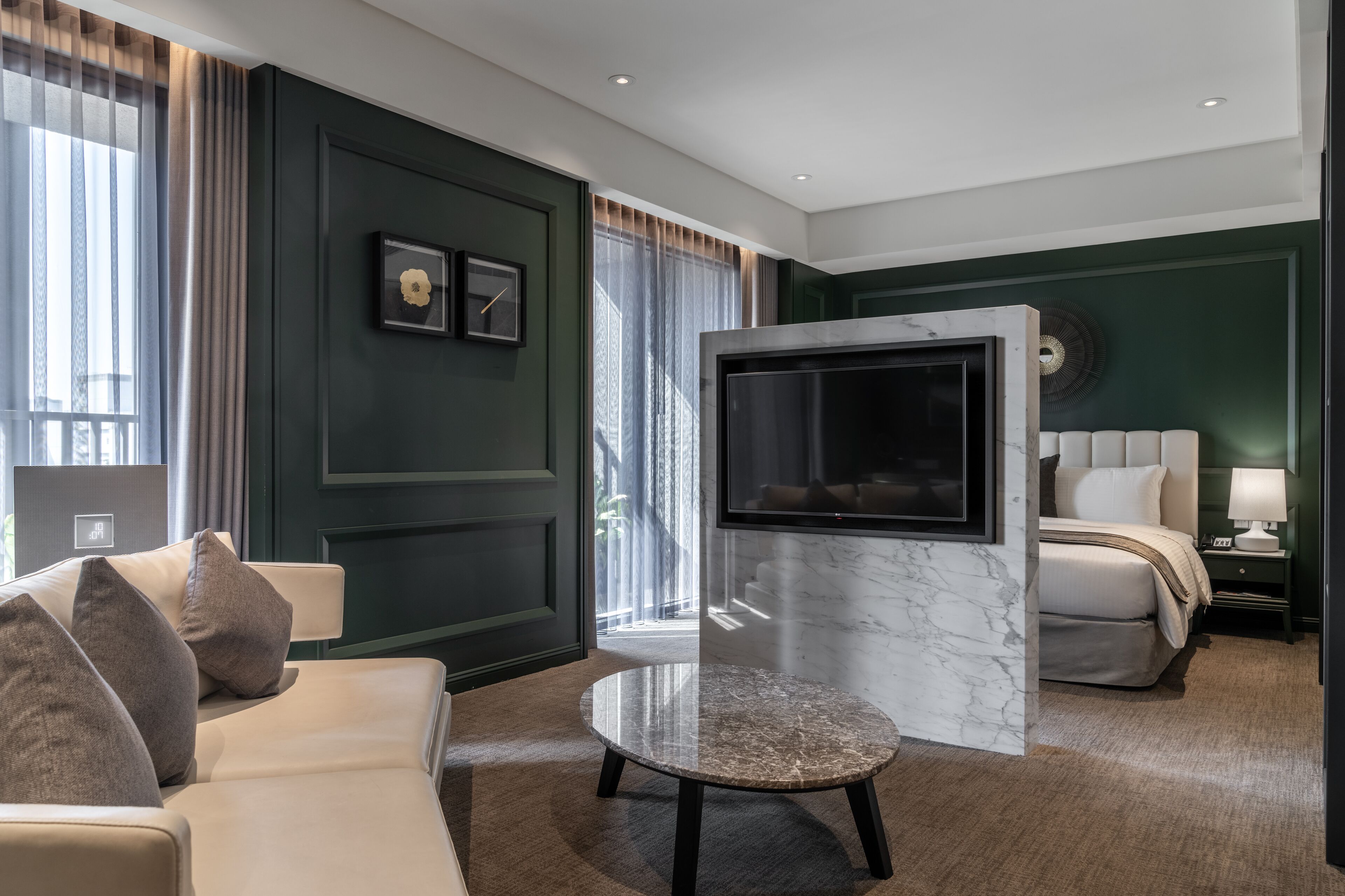 A modern hotel room with a large flat-screen TV, a comfortable sofa, a round coffee table, and a bed with a white headboard.