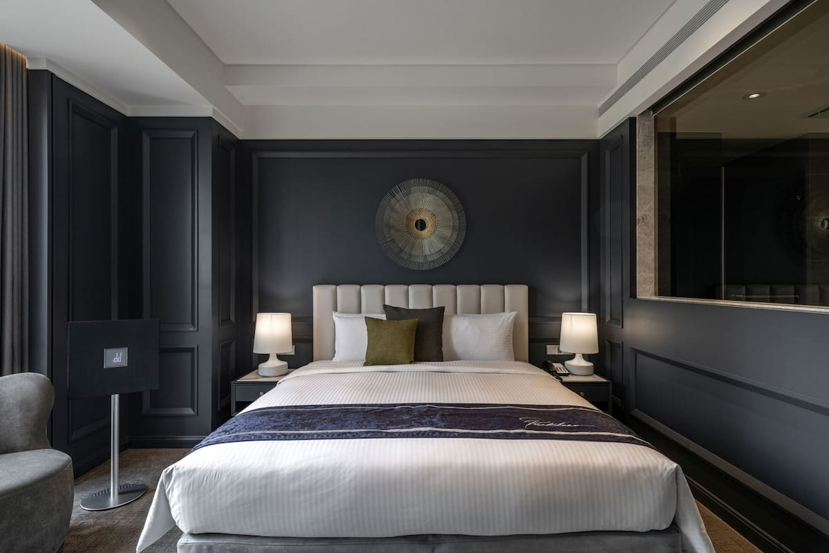 A modern hotel room with a large bed, two bedside tables with lamps, a decorative wall piece, and a large window.