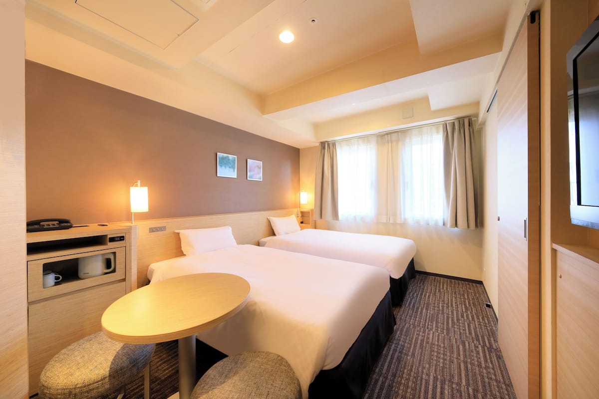 A hotel room with two beds, a small table, a chair, a desk with a lamp, and a TV.