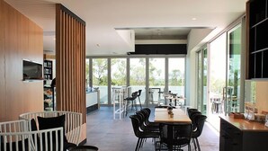 Uma área de refeições moderna com uma mesa comprida e cadeiras, um separador de madeira, e grandes janelas com vista para o exterior.