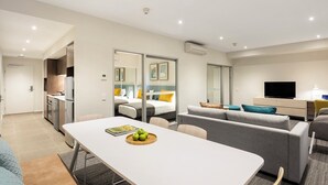 Quarto de hotel moderno com área de refeições, sofá, TV e cozinha compacta.