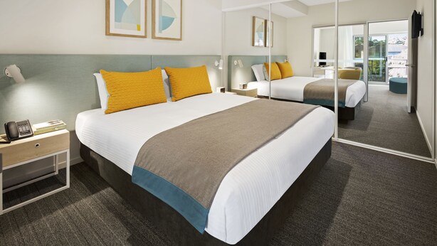 Quarto de hotel moderno com uma cama grande, uma mesa de cabeceira com abajur, um espelho e vista para o exterior.