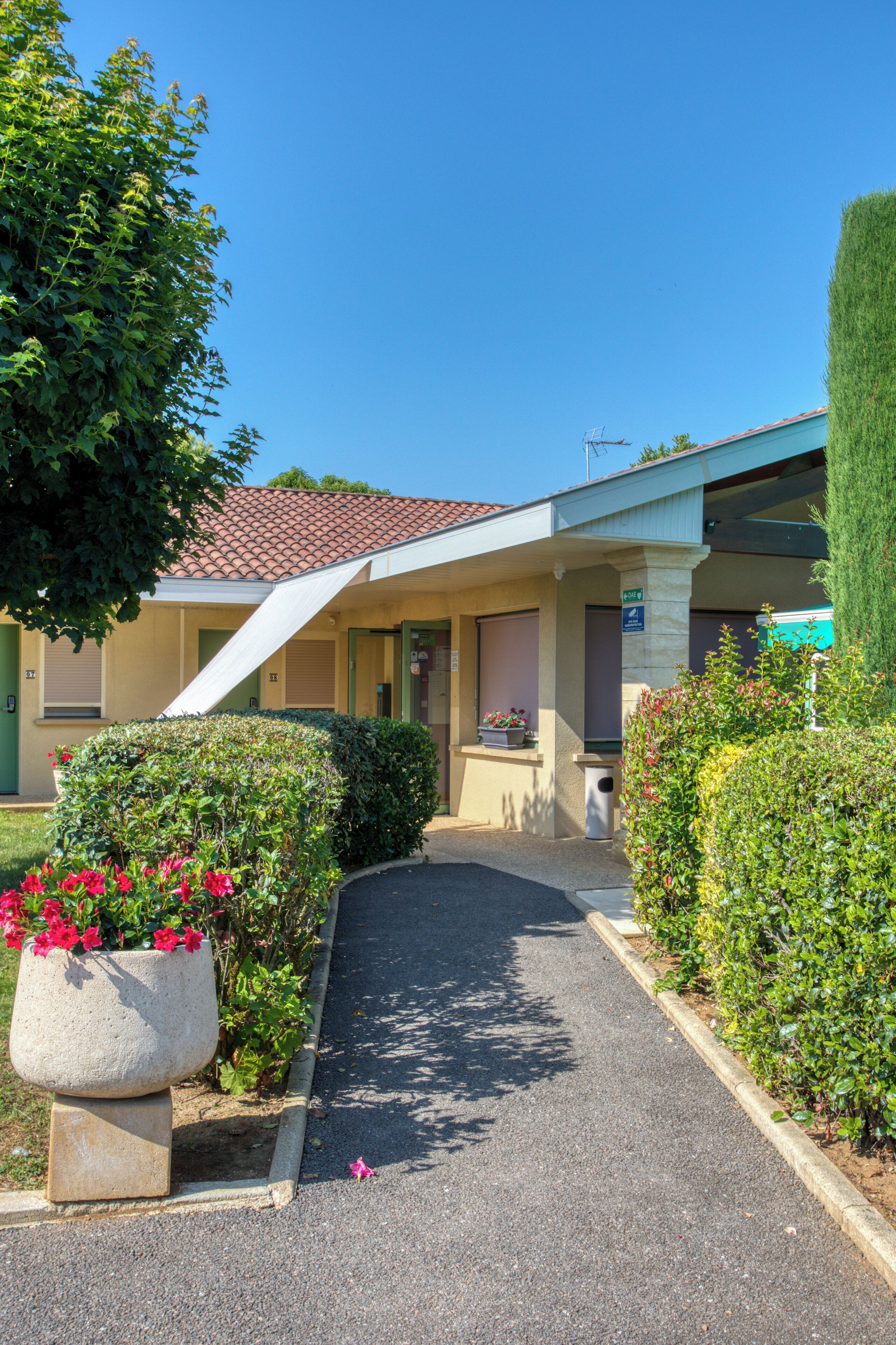 A single-story building with a tiled roof, a covered walkway, and a small garden with a concrete planter and flowers.