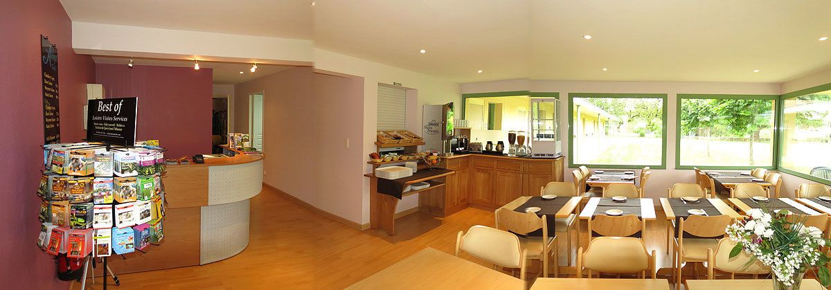 A hotel lobby with a reception desk, a display of magazines, a kitchen area, and a dining area with tables and chairs.