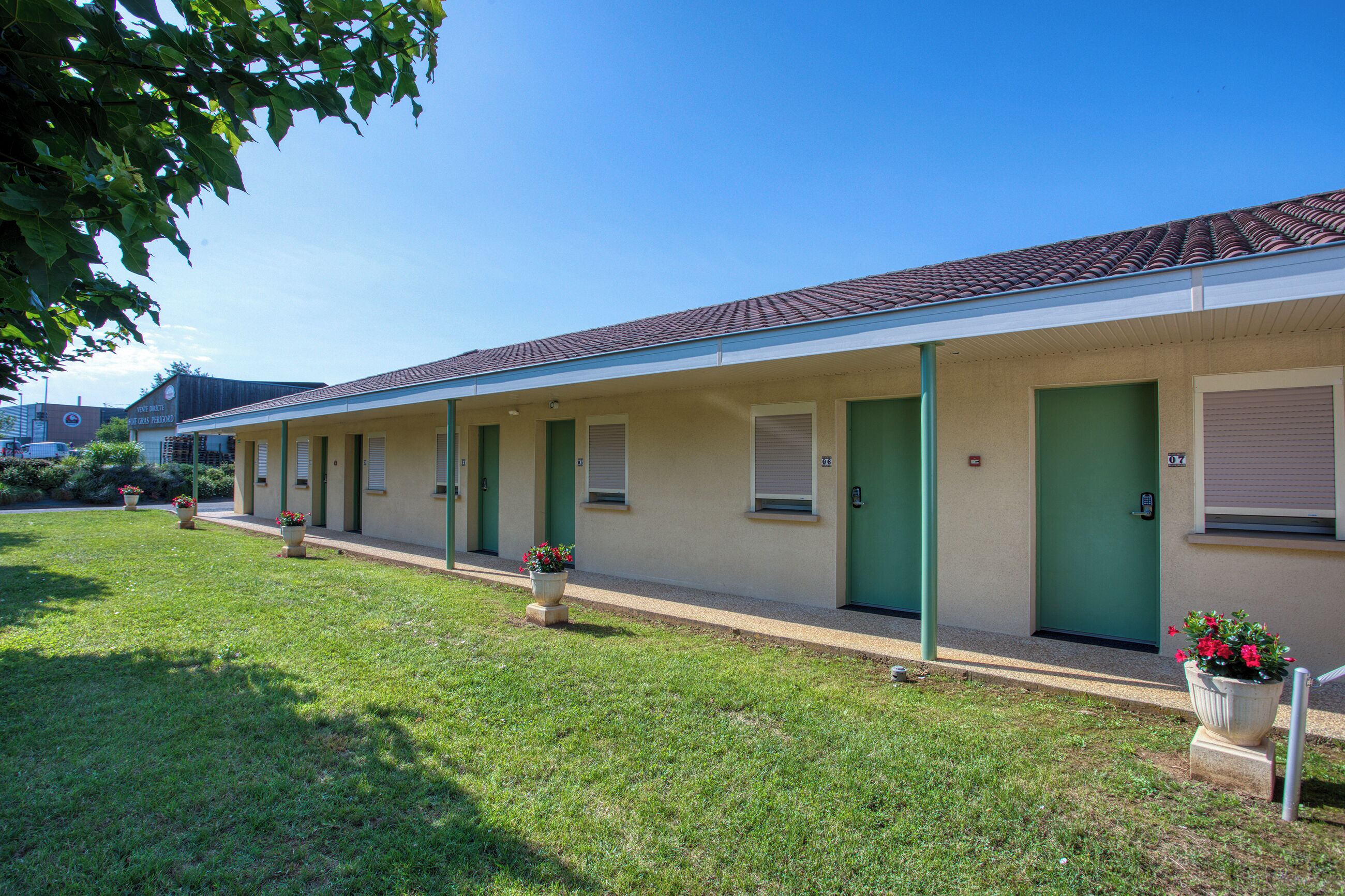 A row of motel rooms with green doors and windows, a covered walkway, and a well-maintained lawn with potted plants.