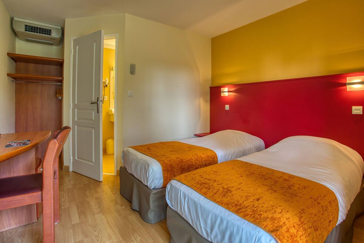 A room with two beds, a desk, a chair, a wardrobe, and a door leading to another room.