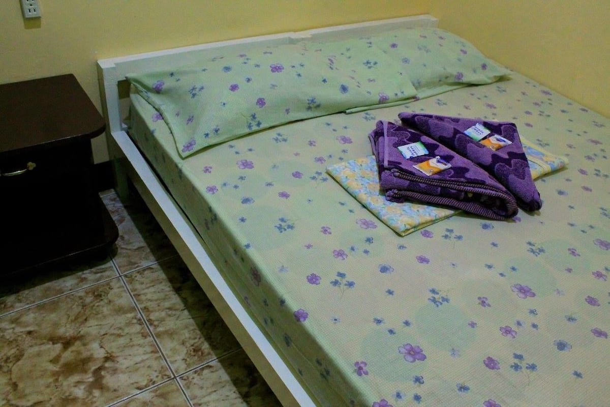A bed with a floral bedspread and pillows, a dark-colored dresser, and a tiled floor.