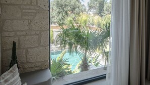 Una stanza con parete in pietra, un'ampia finestra vista piscina e vegetazione, un pouf intrecciato e un divano con una coperta decorata.