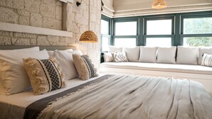 Una camera da letto con parete in pietra, un letto grande con numerosi cuscini, una lampada a sospensione in fibra naturale e vista attraverso una finestra con una panca.