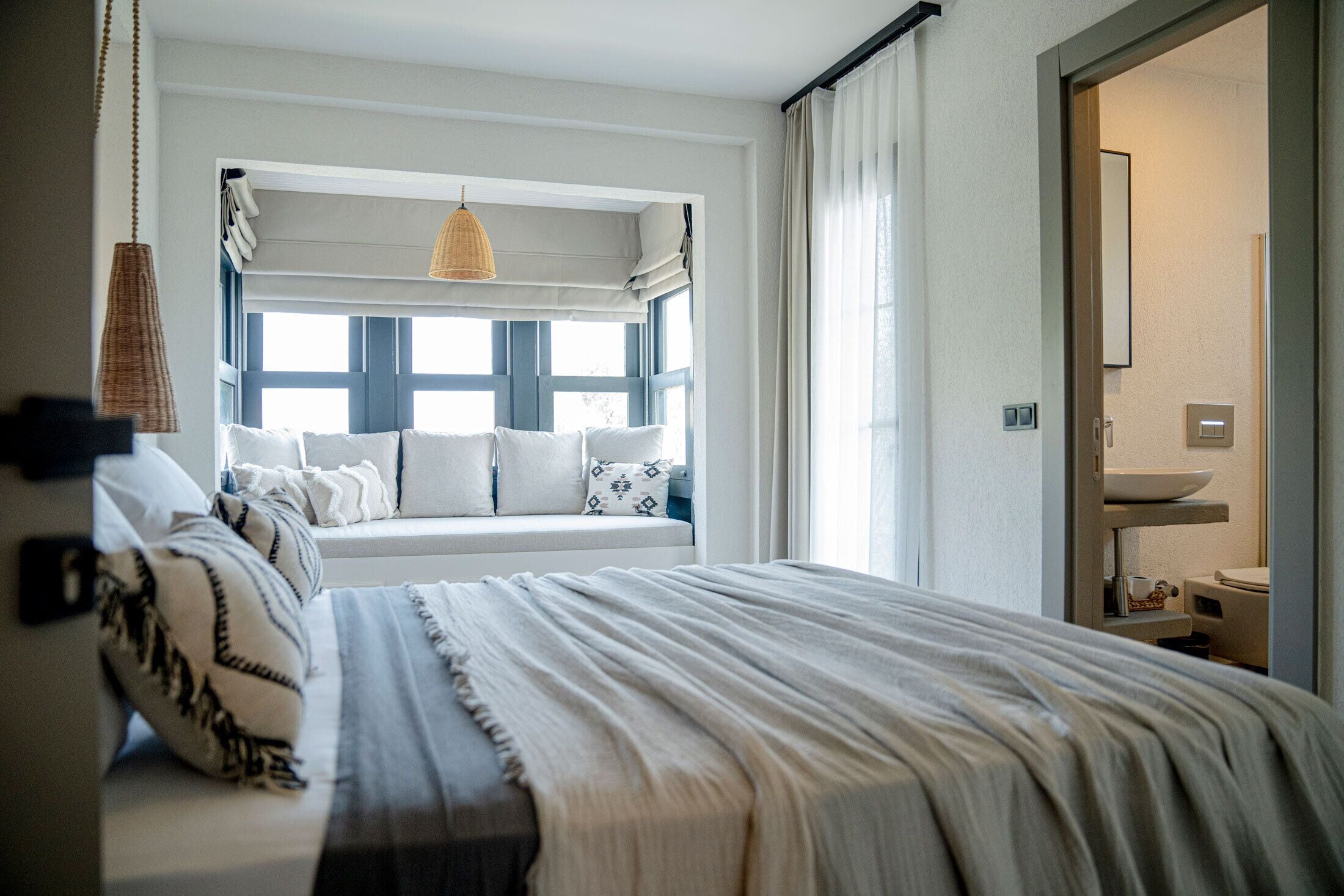 A bedroom with a large bed, a window with blue shutters, a hanging light fixture, and a view of the bathroom.