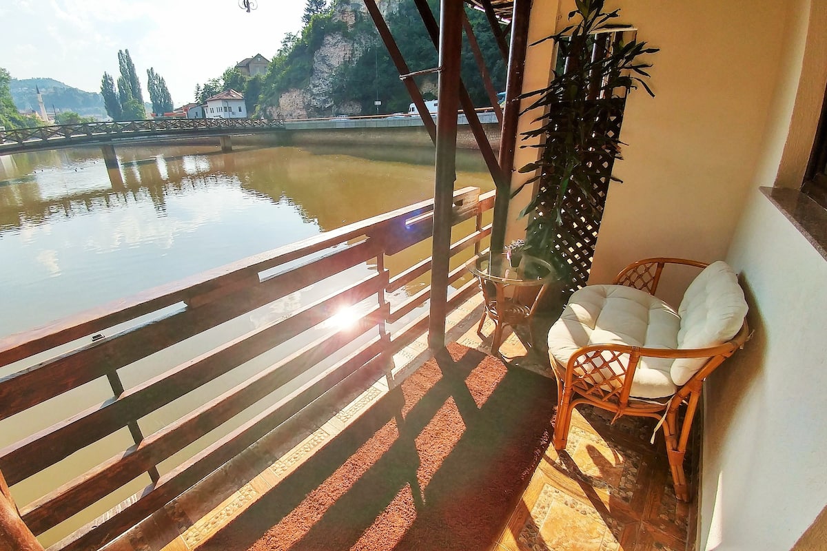 A balcony with a wooden railing overlooking a calm body of water, a wicker chair, and a potted plant.