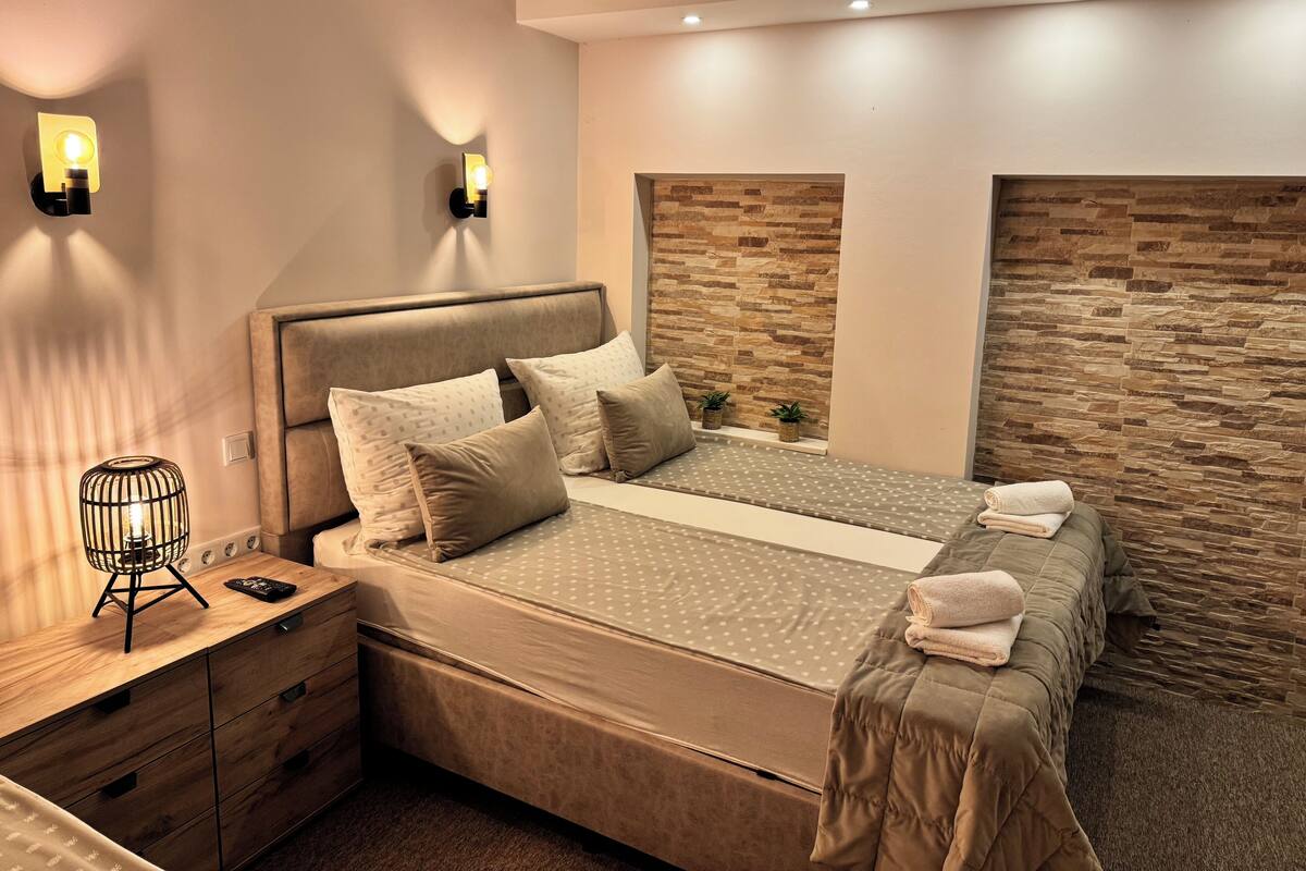 A modern bedroom with a large bed, two wall-mounted lights, a wooden nightstand, and a stone-textured accent wall.