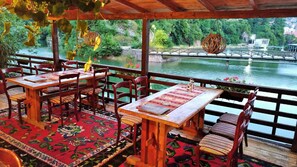 Ein Essbereich im Freien mit Holztischen und -stühlen, einem rot gemusterten Teppich und Blick auf einen Fluss mit einer Brücke.