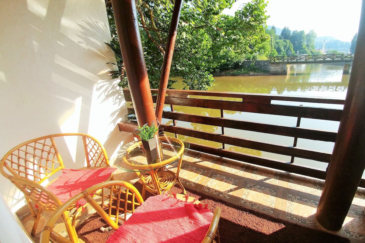 A balcony with a wooden railing, a small table with chairs, and a view of a river and greenery.