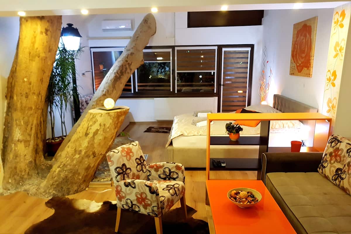 A cozy room with a sofa, a chair, a small table with a bowl, a bed with a headboard, a window with blinds, and a decorative tree trunk.