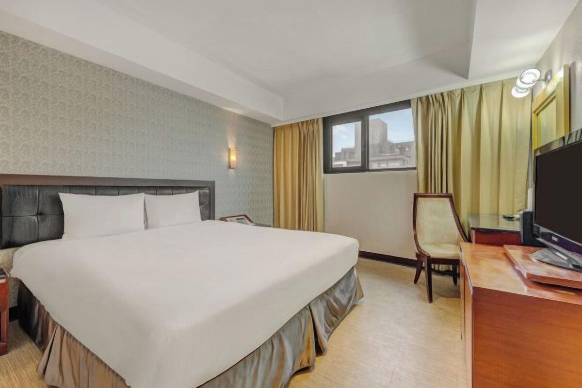 A hotel room with a large bed, a desk, a chair, a television, and a window with curtains.