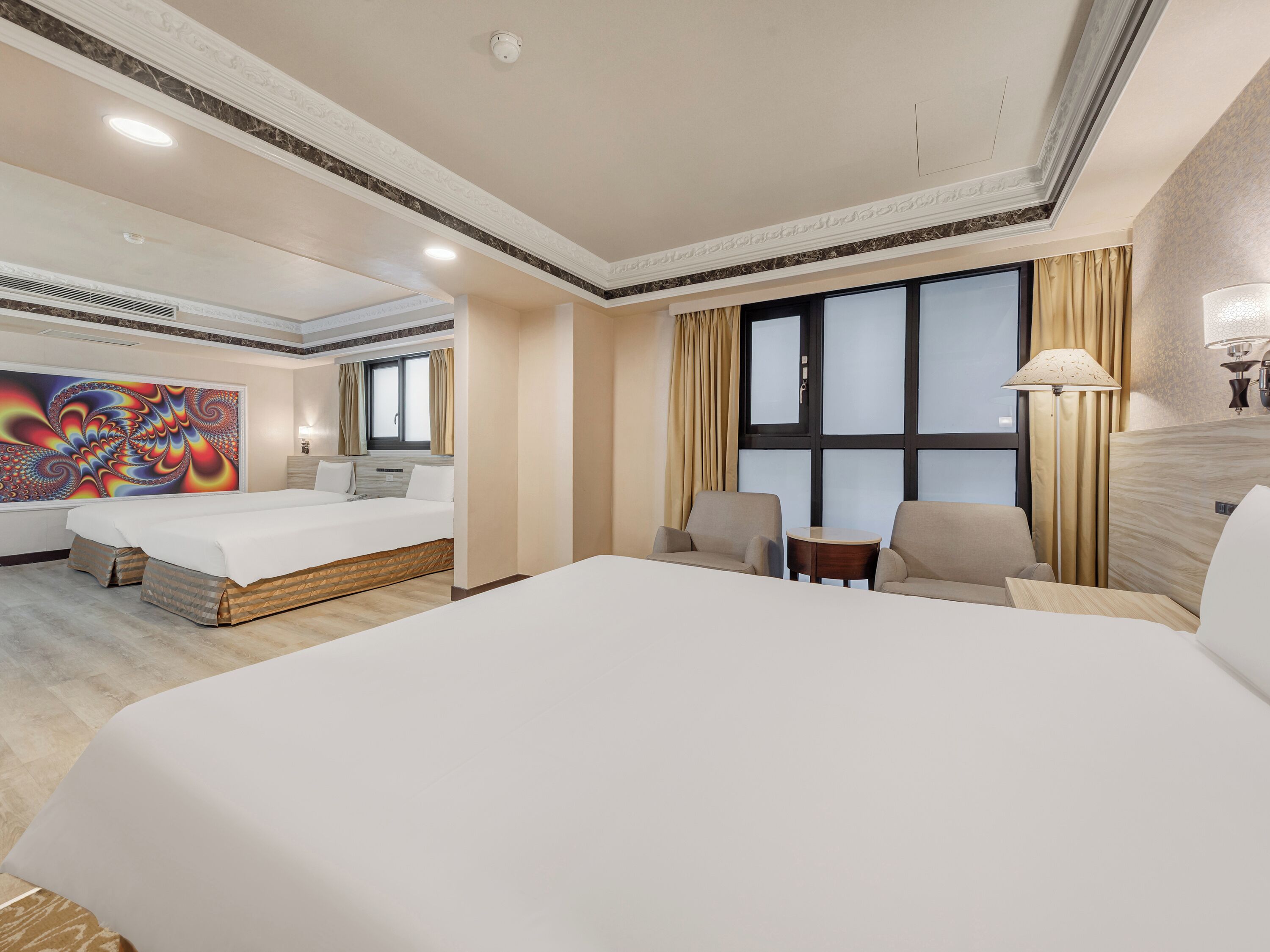 A hotel room with a large bed, two armchairs, a small table, and a colorful artwork on the wall.
