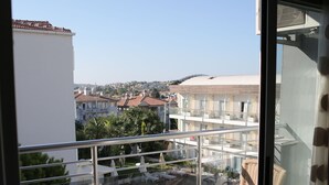 A balcony with wicker chairs, a small round table, and a view of a swimming pool and buildings in the background. - Firat Mert Hotel (Cesme)