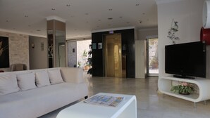 A modern living room with a white sofa, a coffee table with magazines, a flat-screen TV on a white stand, and a large window with a view of the outdoors. - Firat Mert Hotel (Cesme)