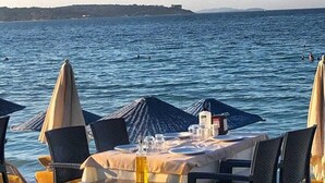 A seaside dining area with tables and chairs, umbrellas, and a view of the sea and distant land. - Firat Mert Hotel (Cesme)