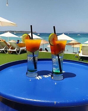 Two drinks on a blue tray with a hashtag symbol, set against a beach background with umbrellas and lounge chairs. - Firat Mert Hotel (Cesme)