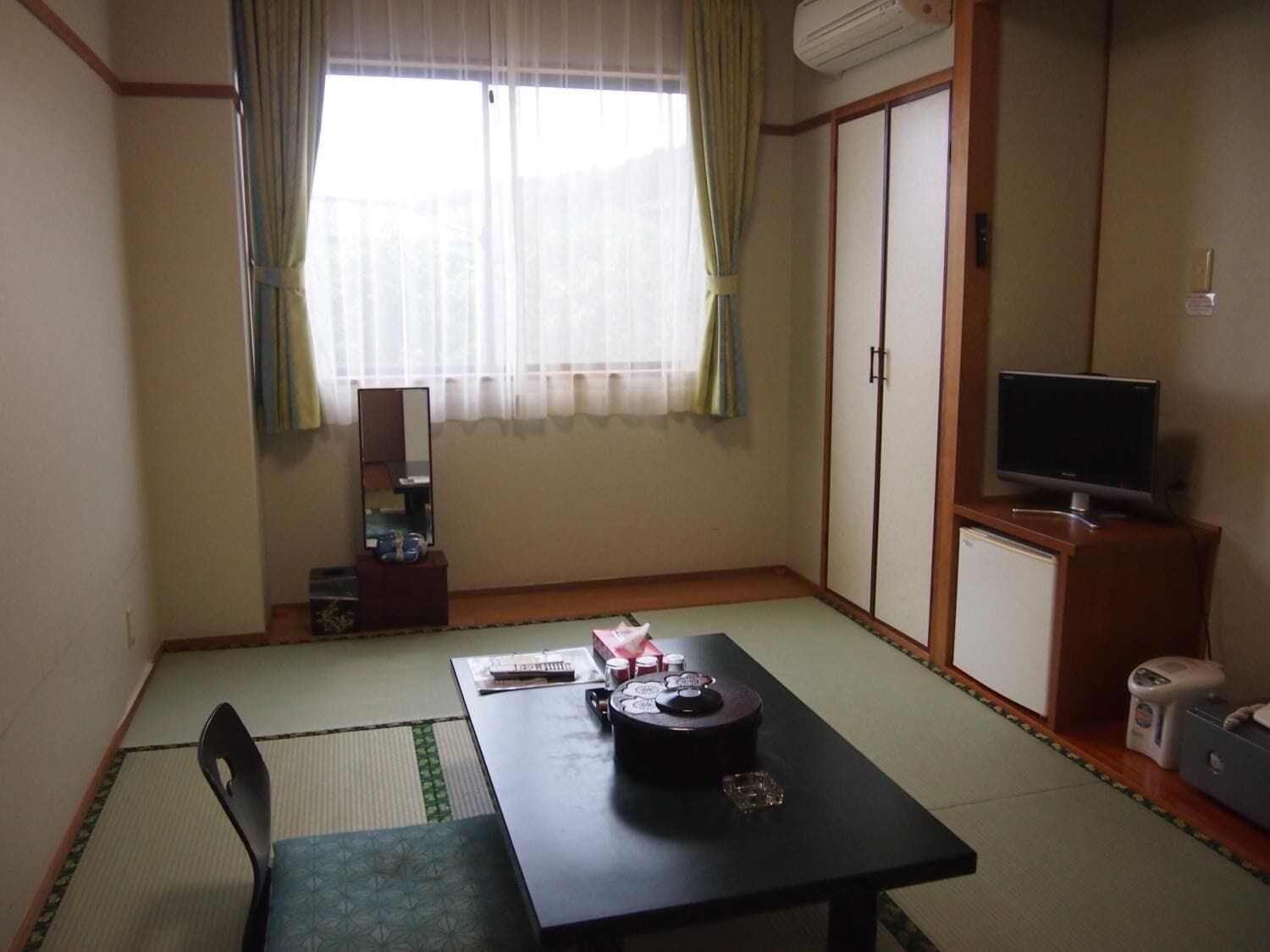 A room with tatami flooring, a low table with a traditional Japanese tea set, a television on a wooden cabinet, and a sliding door closet.