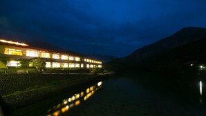 A building with multiple illuminated windows reflecting on a calm water body, surrounded by a stone wall and greenery. - Tashiro Bekkan (Yakushima)