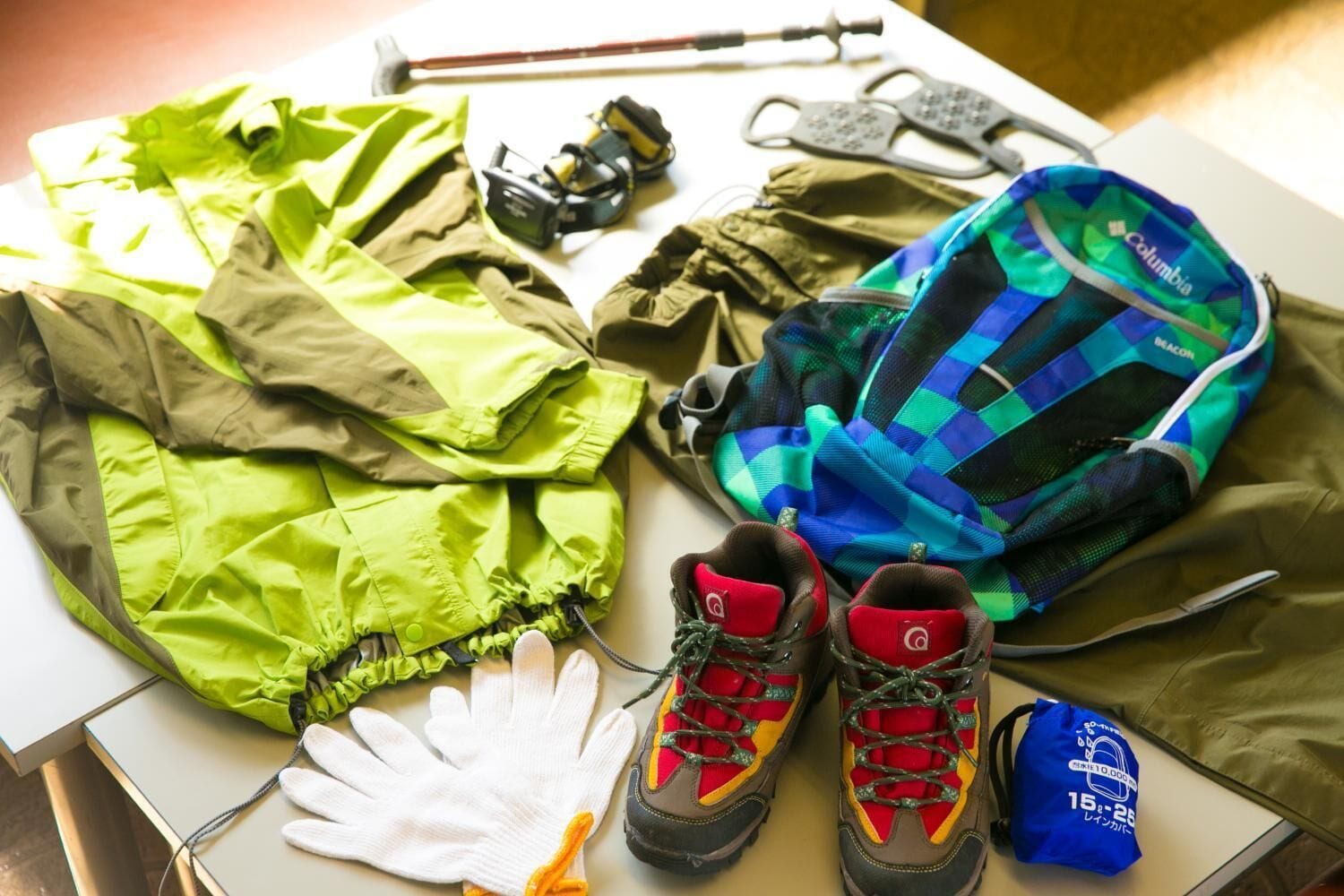 A table with a green jacket, a blue and green backpack, red and black hiking boots, white gloves, and a blue bag with Japanese text.