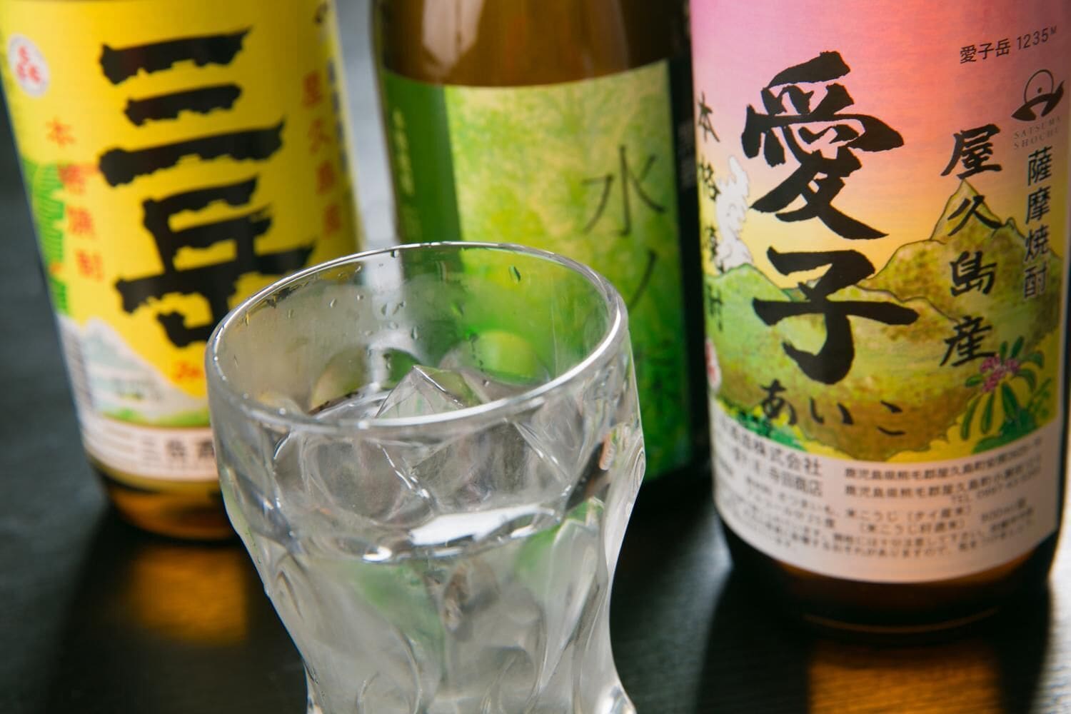 Een glas water met ijsblokjes, twee flessen met Japanse etiketten, en een houten oppervlak.