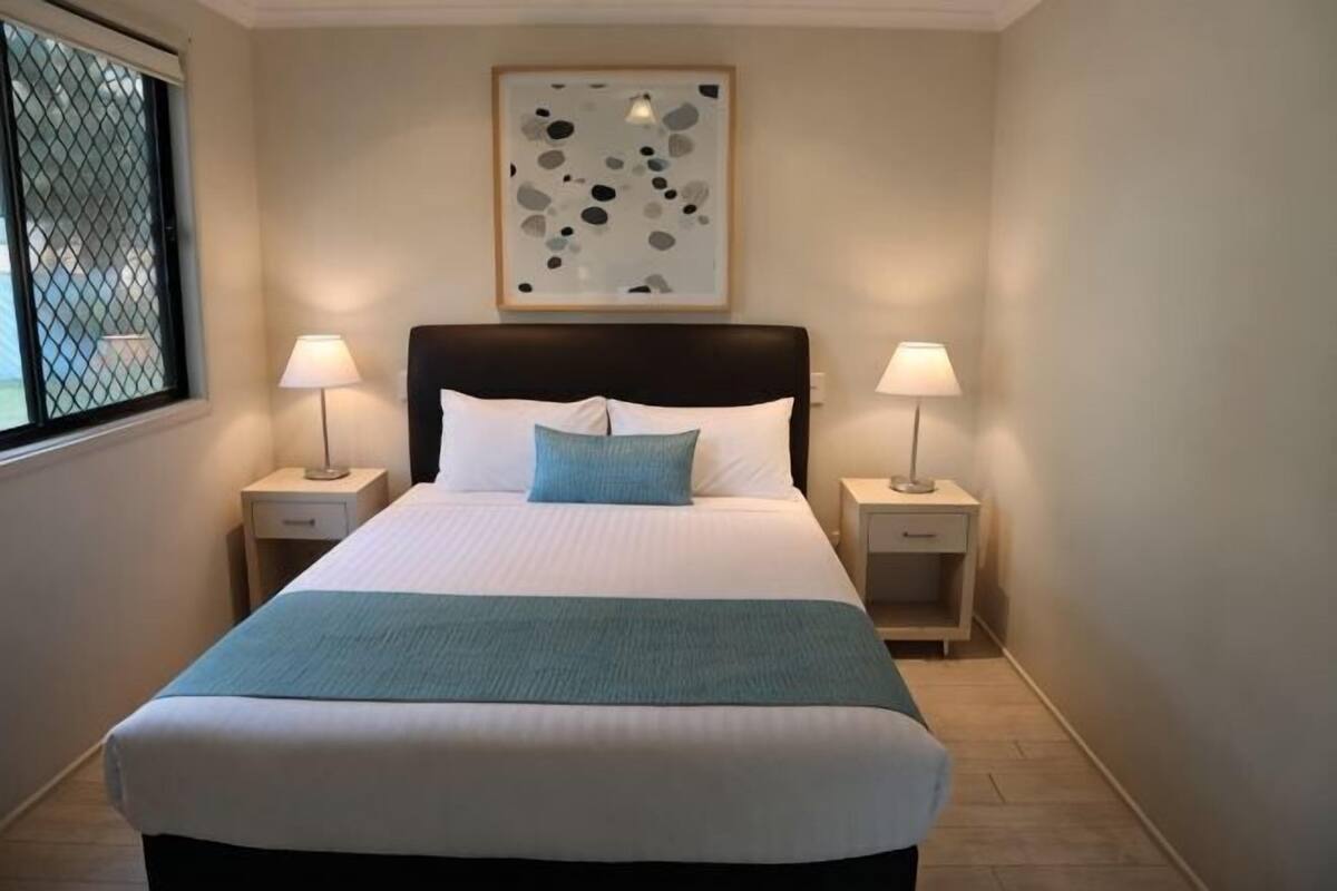 Une chambre d’hôtel avec un grand lit, deux tables de chevet avec des lampes, une fenêtre avec un moustiquaire en métal et une œuvre d’art abstraite accrochée au mur.