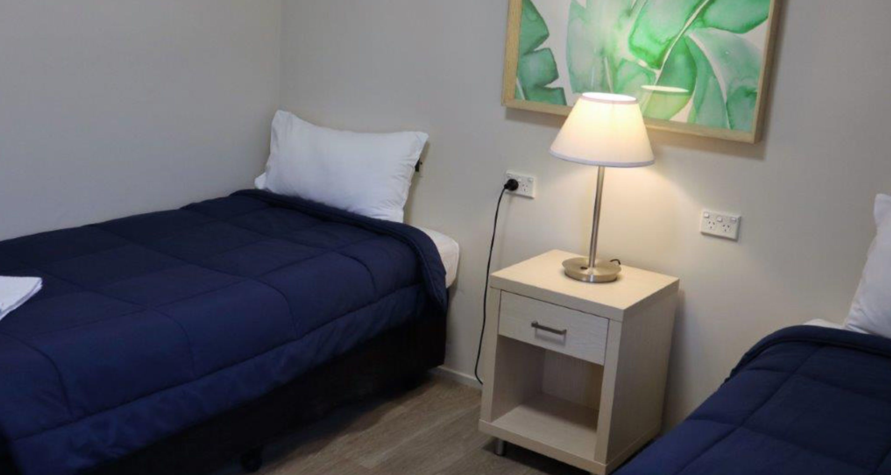 A hotel room with two single beds, a bedside table with a lamp, and a wall-mounted painting.