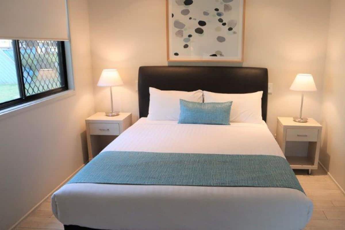 Un lit bien fait, recouvert d’un plaid bleu, avec deux lampes de chevet et, au-dessus du lit, une œuvre d’art abstraite encadrée.