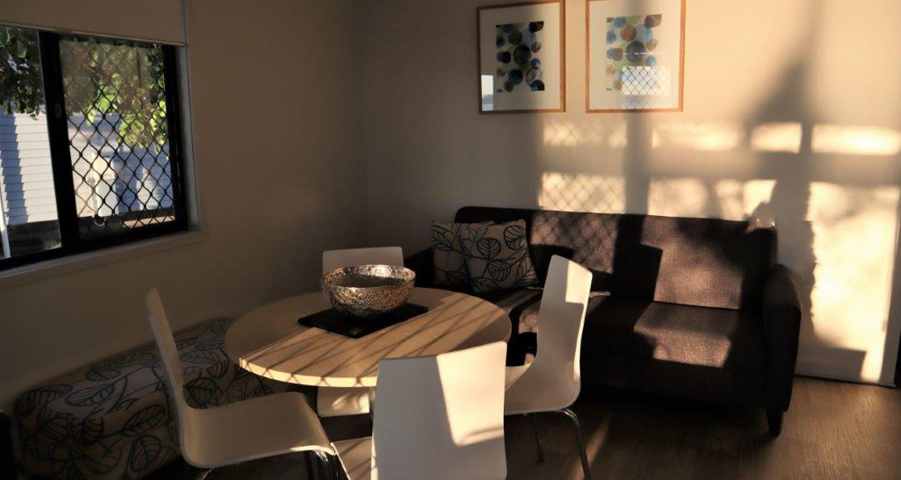 A cozy living room with a round wooden table, white chairs, a patterned sofa, and artwork on the wall.