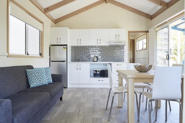 A compact kitchen with white cabinets, a microwave, and a coffee maker. There is a dining area with a table and chairs, and a gray sofa with a patterned pillow.