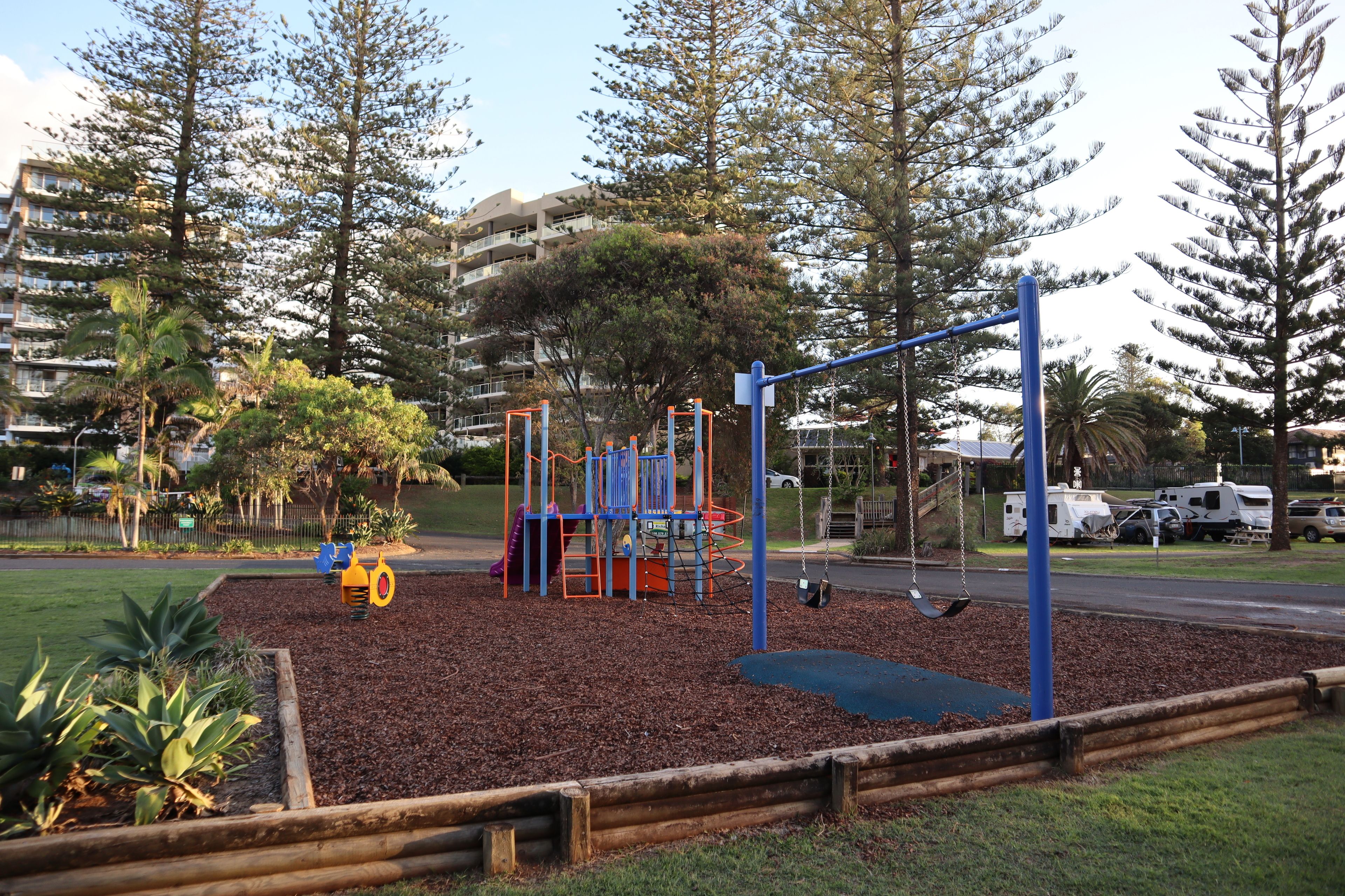 A playground with a swing set and climbing structures, surrounded by a wooden fence and mulch.