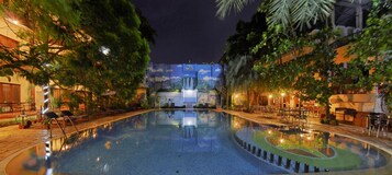 A nighttime swimming pool with lounge chairs and tables, surrounded by trees and buildings with illuminated signs.