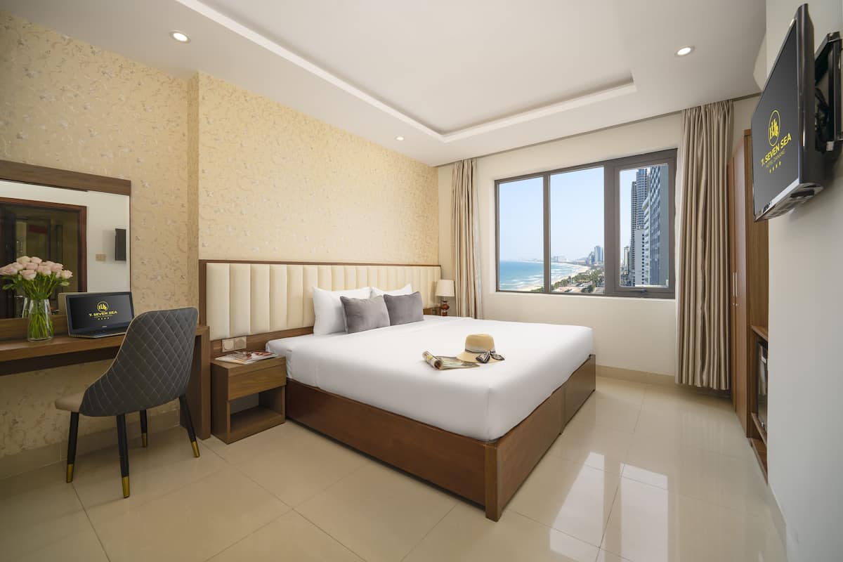 A modern hotel room with a large bed, a desk with a laptop, a chair, and a view of the cityscape through the window.
