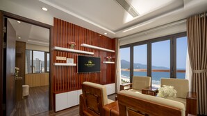 A modern hotel room with a wooden panel wall, a flat-screen TV, a sofa, and a large window offering a view of the sea. - 7 Seven Sea Hotel  (Da Nang)