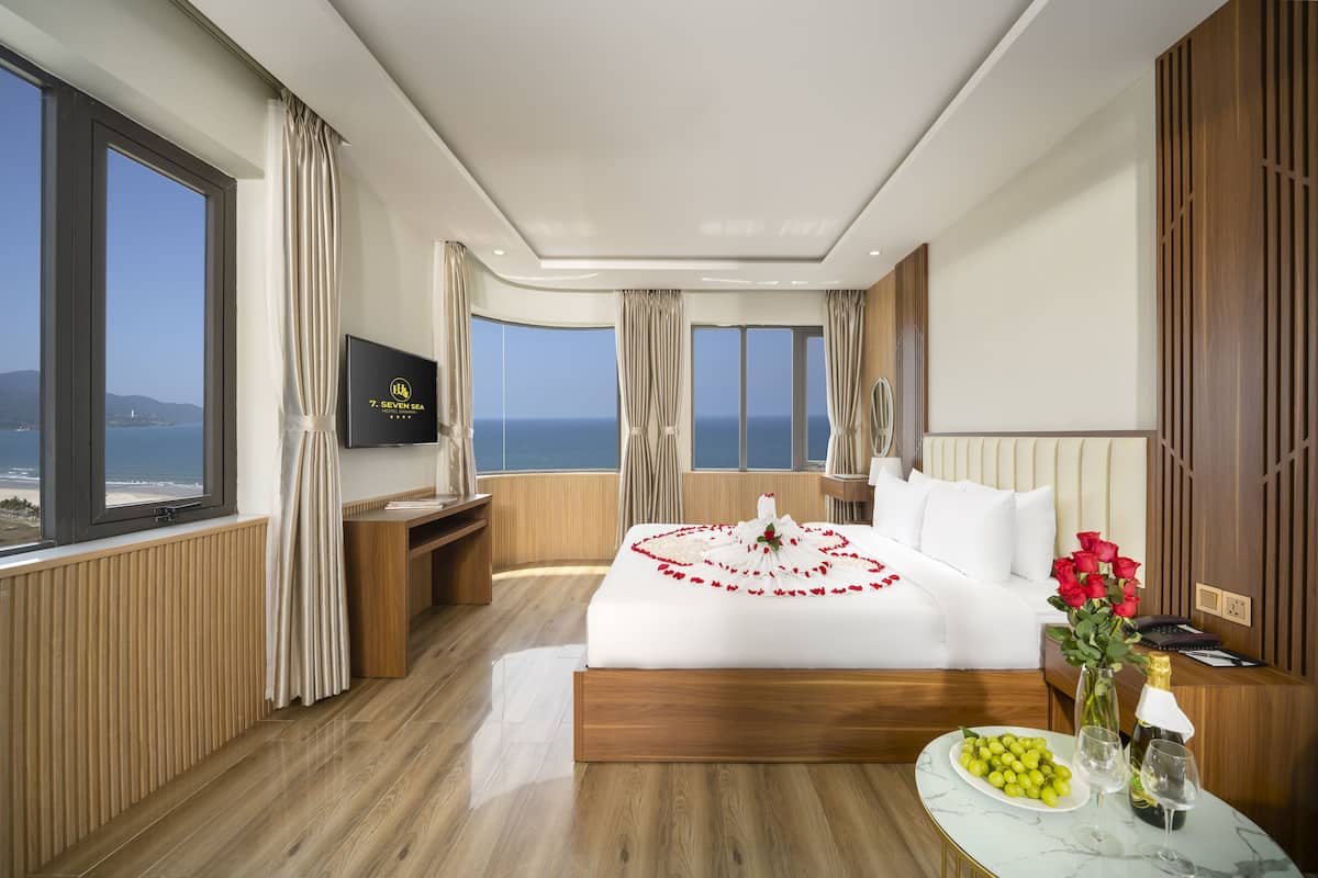 A hotel room with a large bed, a TV, a wooden floor, and a view of the sea.