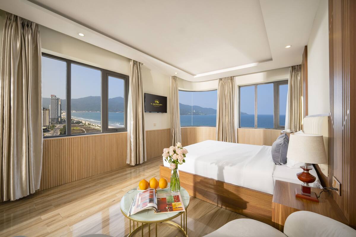 A hotel room with a large bed, a bedside table with a lamp, a small table with a vase of flowers and oranges, and a view of the sea and buildings through large windows.