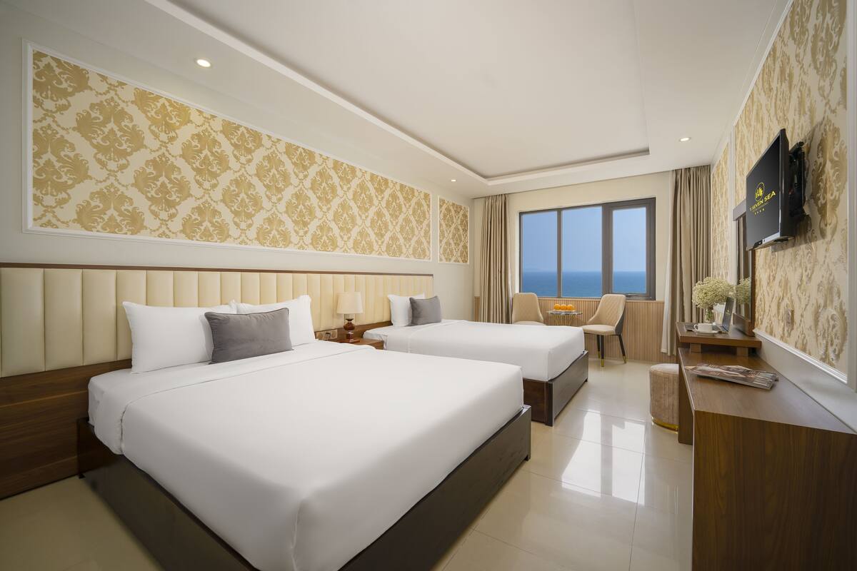 A hotel room with two beds, a desk, a chair, a TV, and a view of the sea.