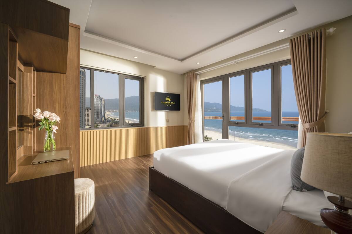 A hotel room with a large bed, a TV mounted on the wall, and a view of the sea through large windows.