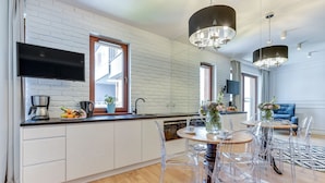 A modern kitchen with a flat-screen TV, a dining area with clear chairs, and a living room with a blue sofa. - GRANO APARTMENTS Gdańsk Motława SPA & Wellness (Gdansk)