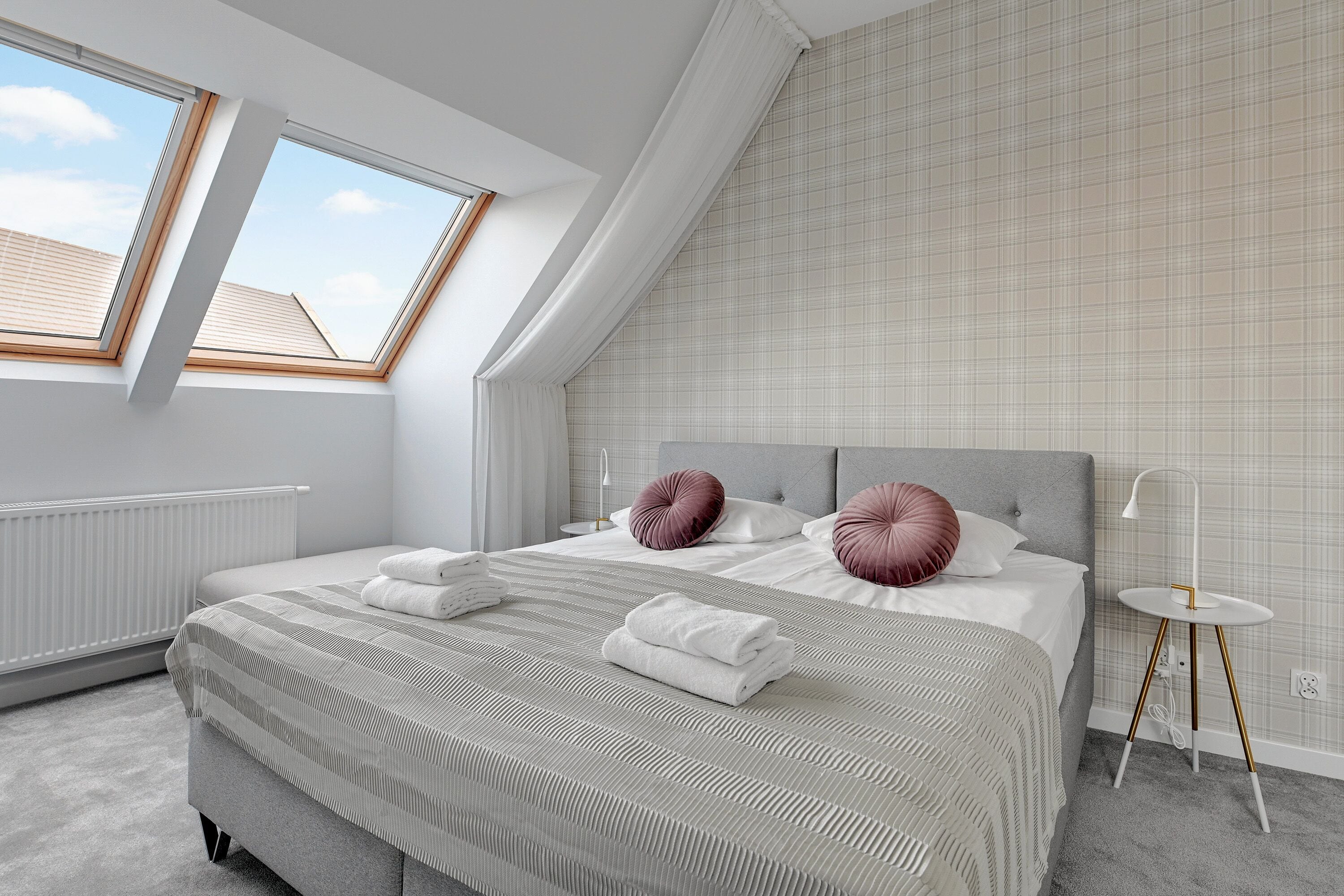 A bedroom with a sloped ceiling, skylights, a bed with pillows, a bedside table, and a radiator.