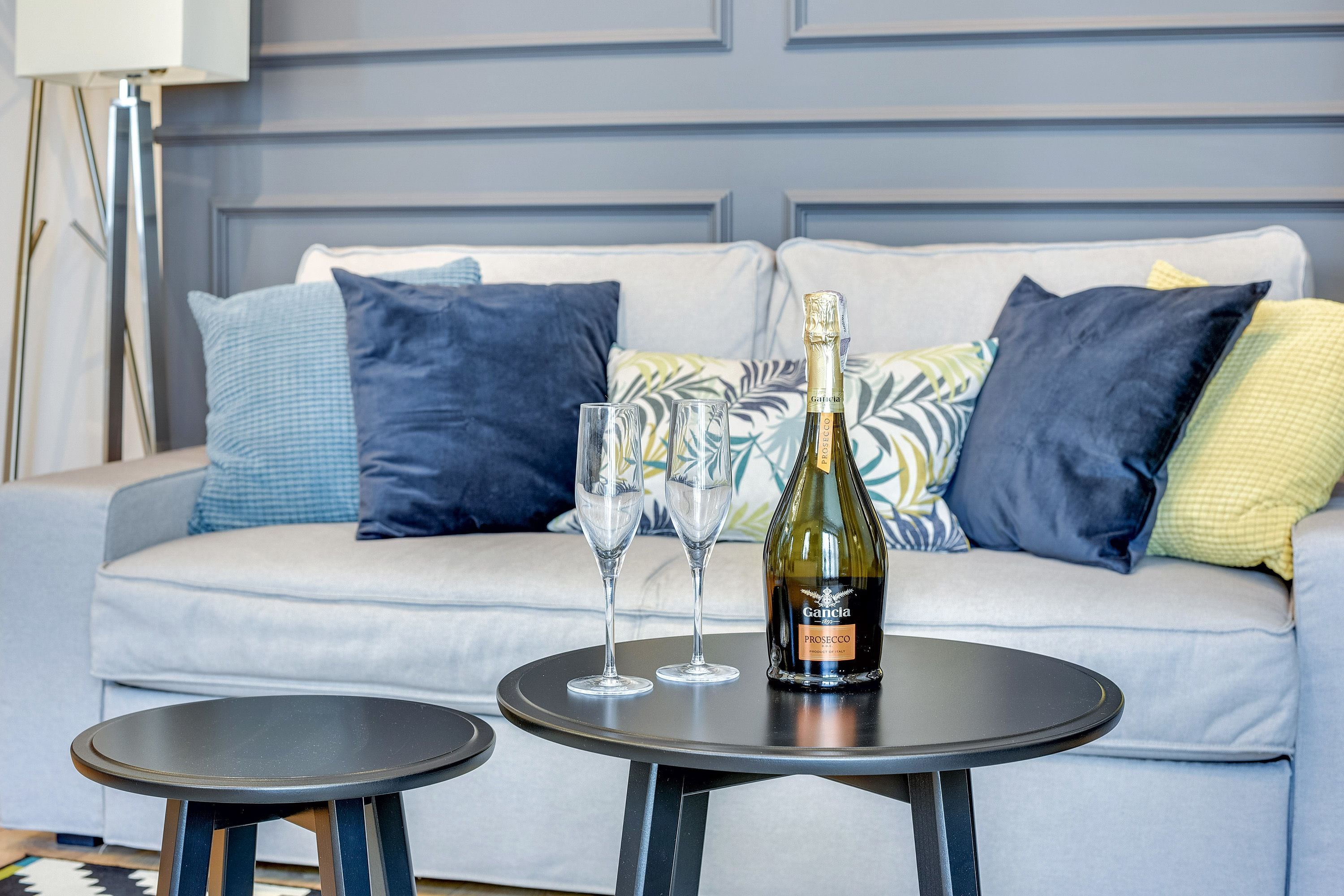 A modern living room with a grey sofa, two small round tables, and a bottle of wine on one table.